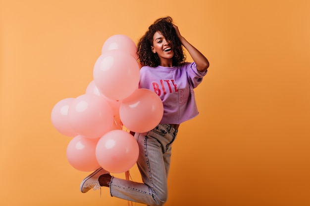 Ecstatic female model in jeans dancing at birthday party. african debonair girl hoding bunch of helium balloons.