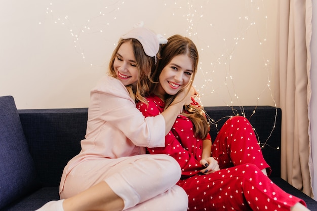 Восторженная девушка в розовой пижаме, сидя на синем софе. Смеющаяся брюнетка в красном ночном костюме позирует, пока ее сестра обнимает ее.