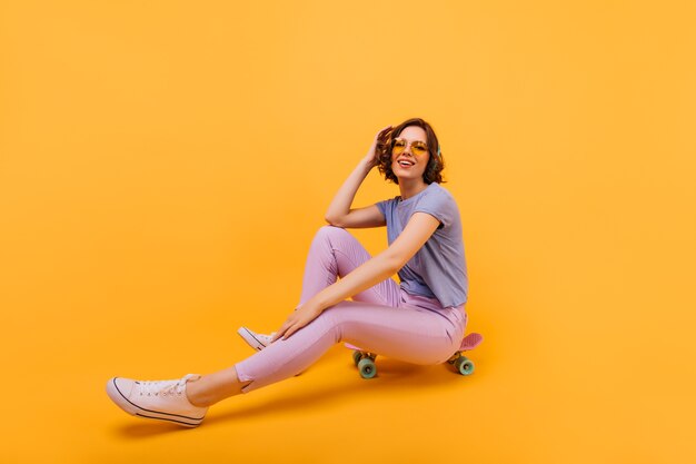ロングボードで良い感情を表現するピンクのパンツの恍惚とした女の子。スケートボードでポーズをとる魅力的な女性モデルの屋内写真。