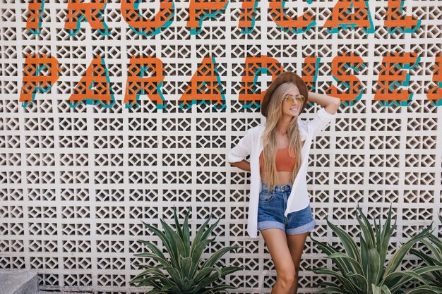 Ecstatic girl with long light hair posing with smile on the street in summer day. Outdoor photo of romantic pretty woman in hat and denim shorts.