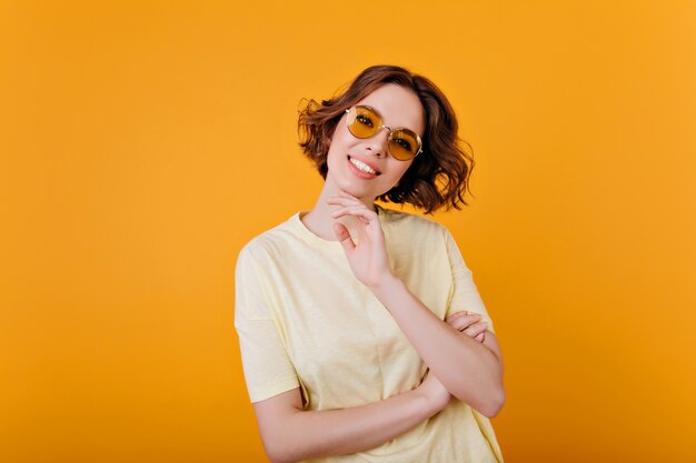 Восторженная бледная девушка в старинных солнцезащитных очках позирует с улыбкой. Фотография в помещении шикарной девушки-модели в светло-желтом наряде.