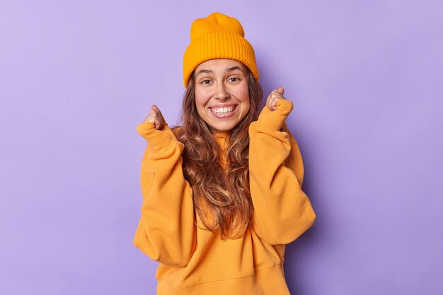 Ecstatic positive woman clenches fists awaits for important results smiles broadly has white teeth feels excited feels enthusiastic and upbeat wears casual loose jumper and hat poses indoor.