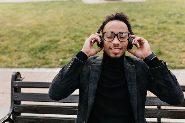 Free photo ecstatic stylish african man in elegant suit sitting on bench. pleased black guy in glasses touching his headphones on lawn