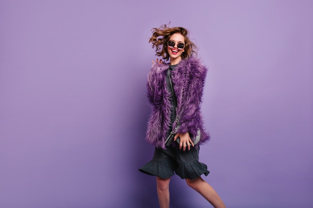 Free photo ecstatic woman in dress and fluffy jacket dancing with laugh