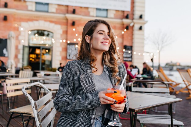 Восторженная женщина в сером пальто с улыбкой смотрит в сторону, попивая фруктовый напиток в кафе