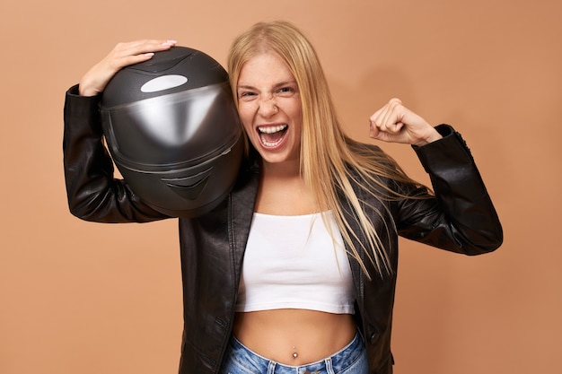 Free photo ecstatic young blonde woman biker in stylish leather jacket keeping fist clenched and mouth wide opened, screaming