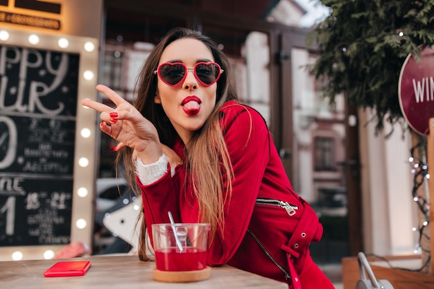 Ecstatic young female model in red glasses posing with tongue out