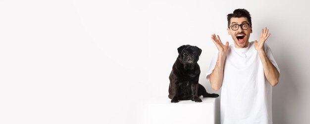 Ecstatic young man looking with excitement and rejoice standing near cute black pug staring at camer