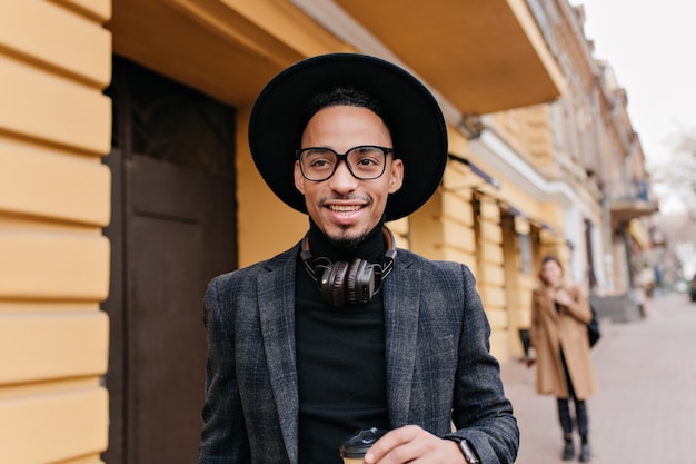 Free photo ecstatic young man with brown skin looking away with dreamy smile. outdoor portrait of handsome smiling black guy standing near restaurant.