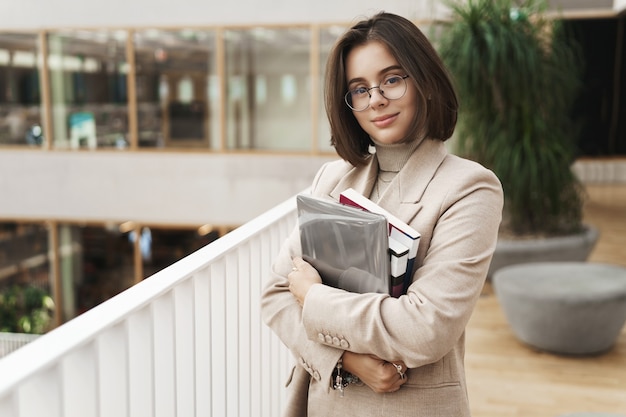 Foto gratuita concetto di istruzione, affari e donne. ritratto di giovane tutor femminile attraente, elegante, giovane insegnante o studente porta a studiare libri e laptop, in piedi nella fotocamera sorridente del corridoio.