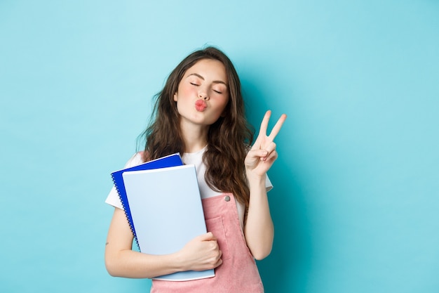 Education concept. Smiling glamour girl pucker lips for kiss, showing v-sign peace and holding notebooks for study, standing against blue background