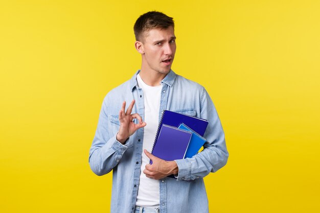 Education, courses and university concept. Handsome assured blond guy encourage enroll this college, showing okay gesture as guarantee you will like it, holding notebooks, yellow background.