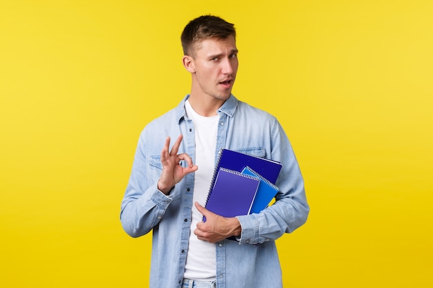 Education, courses and university concept. Handsome assured blond guy encourage enroll this college, showing okay gesture as guarantee you will like it, holding notebooks, yellow background.