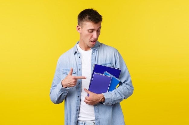 Education, courses and university concept. Handsome young male student pointing finger at notebooks and study material, prepare homework for college assignment, yellow background.