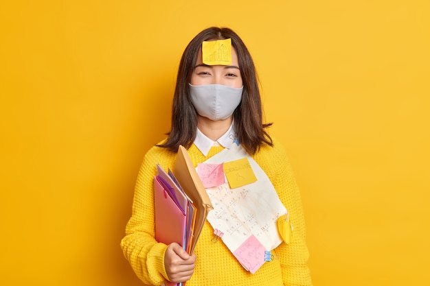 Education social distancing and self isolation concept. Asian unversity student wears protective face mask during coronavirus carries folders and memo stickers prepares for final exam from home