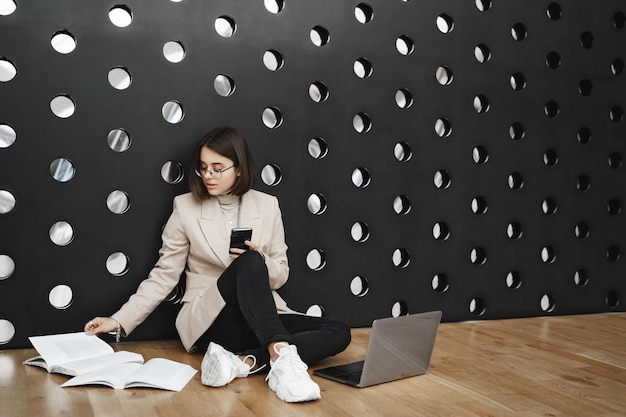 Free photo education student lifestyle and career concept portrait of young smart girl working on project sit coworking space or college lounge on floor reading book and type report in laptop