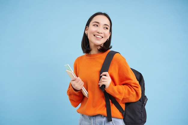 Istruzione e studenti sorridente giovane donna asiatica con zaino e quaderni in posa contro il bac blu