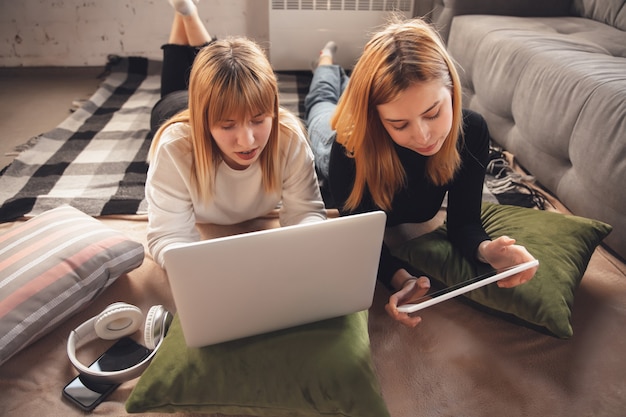 Education time. Young friends, women using gadgets to watch cinema, photos, online courses, taking selfie or vlog, shopping. Two caucasian girls at home using laptop, tablet, smartphone, headphones.