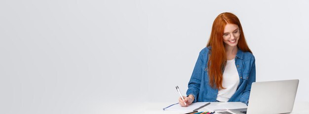 Free photo education work and vanlife concept cheerful lovely redhead woman in glasses standing near table read