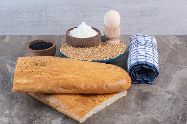 Egg cup, flour bowl and wheat pile on a platter next to black sesame bowl, roll of towel, and bread loaves on marble background. High quality photo