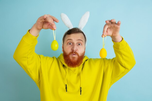 Egg hunt coming. Caucasian man as an Easter bunny with bright casual clothes on blue studio background. Happy easter greetings. Concept of human emotions, facial expression, holidays. Copyspace.