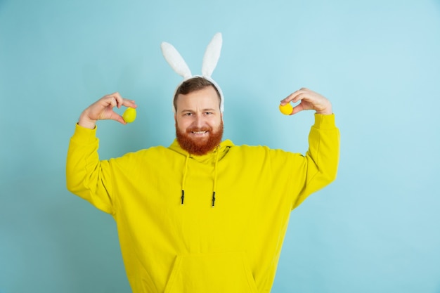 Egg hunt coming. Caucasian man as an Easter bunny with bright casual clothes on blue studio background.