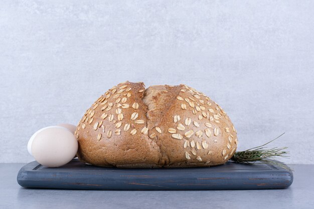 An egg, a loaf of bread and a single wheat stalk on a board on marble surface