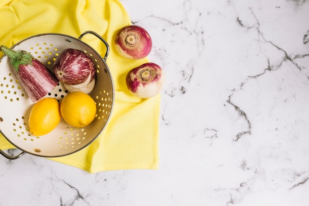 Eggplant; turnip and lemons in colander over yellow napkin on white marble background