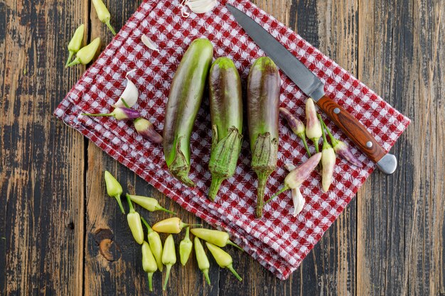 Free photo eggplants with garlic, peppers, knife on wooden and kitchen towel, flat lay.