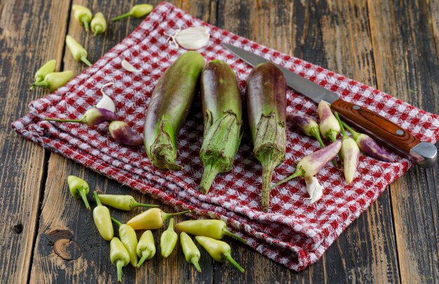 Eggplants with garlic, peppers, knife on wooden and kitchen towel, high angle view.