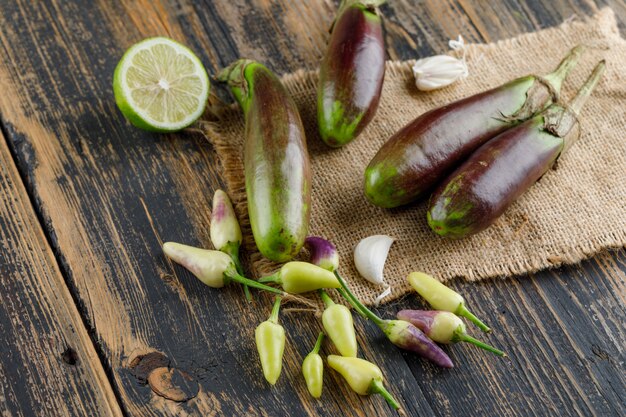 Eggplants with lime, garlic, peppers on wooden and piece of sack, high angle view.