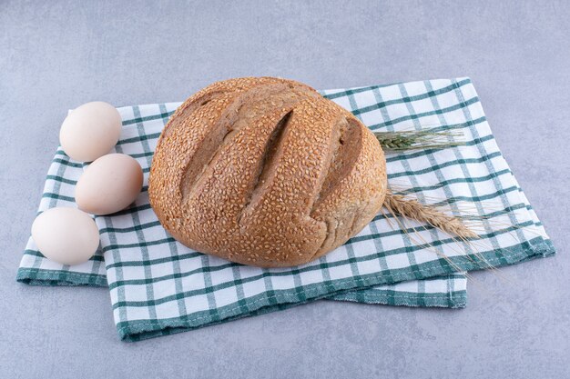 Eggs, bread loaf, wheat stalks placed on a folded towel on marble surface