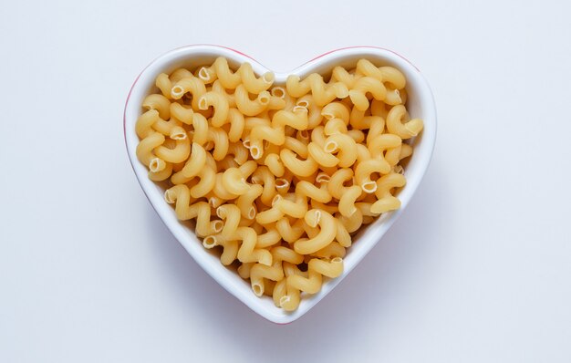Elbow macaroni pasta in a heart shaped bowl top view on a white table