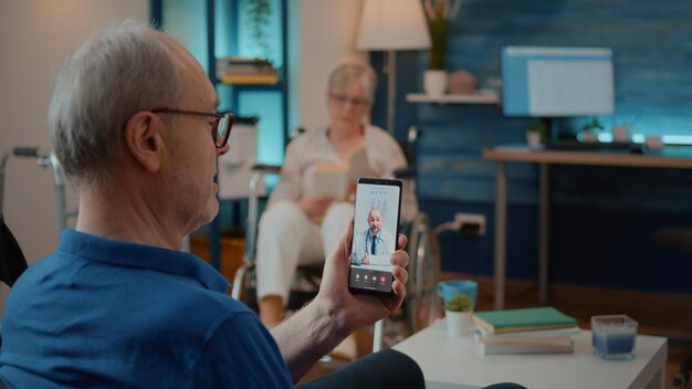 ビデオ通話を使用して自宅での治療について医師に相談する高齢者。遠隔医療および遠隔医療のための遠隔電話会議のヘルスケアについて専門家と話している年配の男性。
