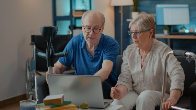 Elder people with walking frame and crutches using laptop in living room. Retired man and woman with chronic disability browsing internet on computer and enjoying technology on free time.