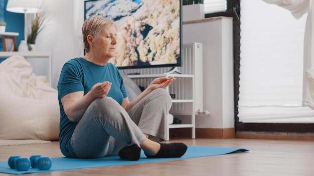 Elder person sitting in lotus position on yoga mat to meditate at home. Senior woman doing zen meditation for calm and balance, relaxing after fitness training. Pensioner meditating.