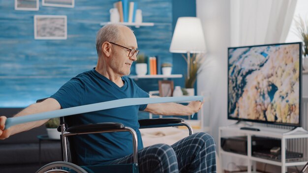 Elder person with disability stretching resistance band to exercise, looking at training lesson on tablet. Senior man sitting in wheelchair and pulling elastic belt to train with workout video