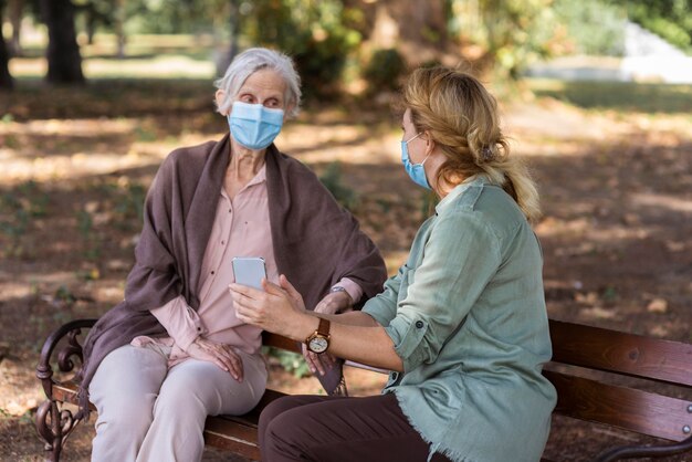 Elder woman conversing with woman on bench outdoors while holding smartphone