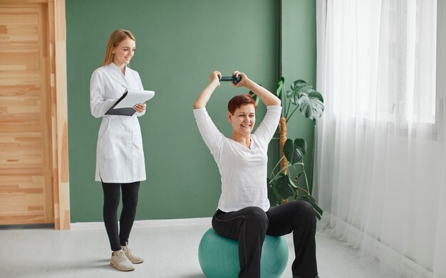 Elder woman in covid recovery doing physical exercises with dumbbell while nurse checks