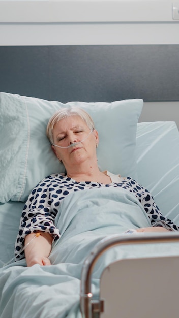 Free photo elder woman with iv drip bag sleeping in hospital ward bed while nurse checking heart rate monitor for healthcare and recovering. sick patient resting with nasal oxygen tube for recovery.