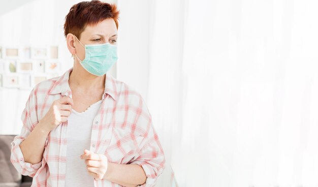 Elder woman with medical mask looking through window at home