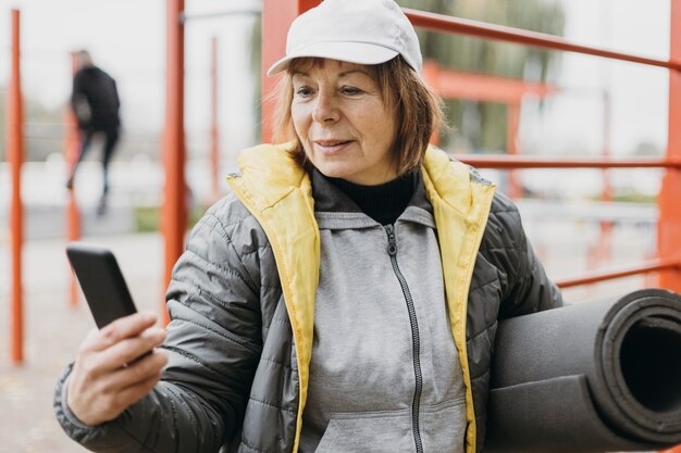 Elder woman working out outdoors while holding smartphone and mat