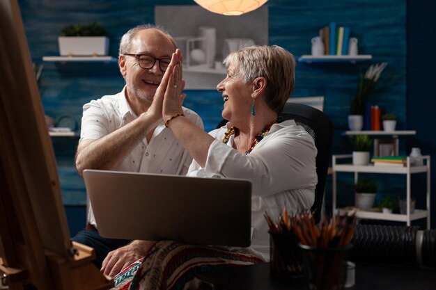 Elderly artist couple holding laptop doing high five hand gesture celebrating selling artwork in online auction sitting in home studio. Senior art creators enjoying success on social media.