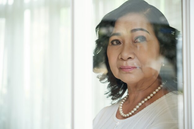 Elderly Asian lady looking outside through window glass