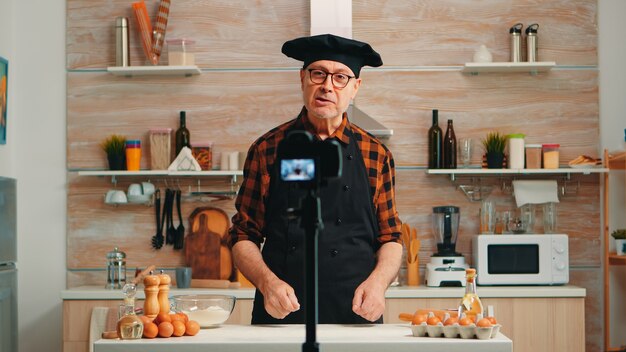 Elderly baker recording video tutorial about food recipe in kitchen. Retired blogger chefinfluencer using internet technology communicating, shooting, blogging on social media with digital equipment