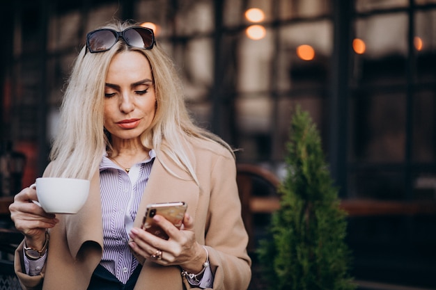 Elderly businesswoman drinking coffee outside cafe and talking on the phone