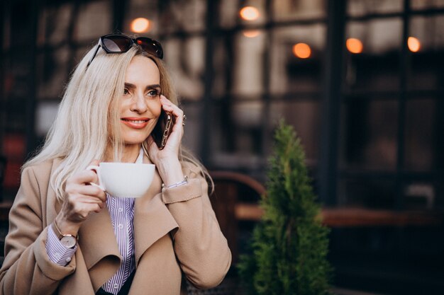 Elderly businesswoman drinking coffee outside cafe and talking on the phone