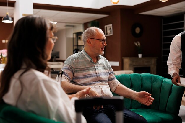 Free photo elderly couple on comfortable sofas receiving assistance from helpful resort employee retired senior man receiving cup of coffee from african american waiter while talking to his wife