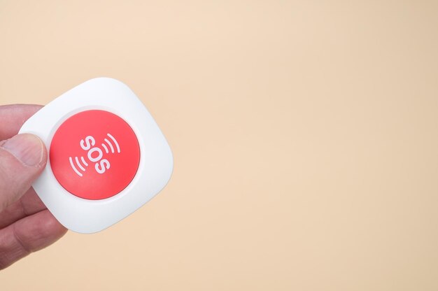 明るい茶色の背景に分離されたワイヤレスSOS緊急警報ボタンを持っている高齢者の手