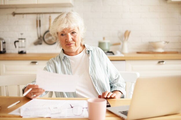 開いたラップトップとテーブルの上に書類を置いてキッチンに座っている白髪の年配の主婦は、感情的な欲求不満の表情をしており、国内の請求書をオンラインで支払っている間、借金の金額にショックを受けました